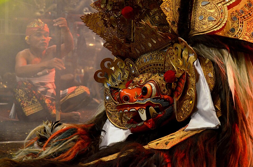 Barong Bali — the sacred lion-spirit costume of Bali's Barong dance tradition, showing the elaborate ornate golden mask with bulging eyes and fangs, the thick white-and-gold fur body, floral decorations, and mirror ornaments that characterise Barong Ket — the most widespread form of the Barong, representing the King of Spirits and the embodiment of Dharma (cosmic good), whose costume is treated as a sacred living object rather than theatrical dress, directly parallel to Theyyam's sacred headdress (Mudi)