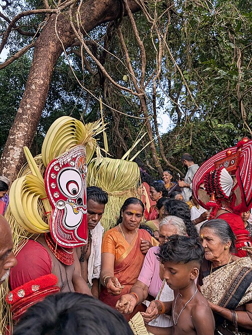 Gulikan Theyyam at Arayi Karthika Kavu, North Malabar — close view of the performer's Mukhathezhuthu face painting in red, white, yellow and black natural pigments, the trishul mark of Shiva on the mask's forehead, and the costume of coconut leaves and bamboo slivers adorned with Katakam and Chutakam ornaments, demonstrating the elaborate transformation of the performer into the deity of death and destiny