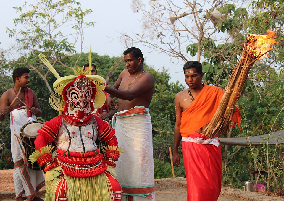 Gulikan Theyyam Purappad at Kannapuram, North Malabar — the performer in full Kolam costume with the towering areca-palm Mudi headgear shaped like a ladder, codified red Mukhathezhuthu face paint mixed from turmeric and limestone, and the trishul mark of Shiva on the mask's forehead, emerging in procession during a Kaliyattam festival to embody the fierce guardian deity of death and destiny