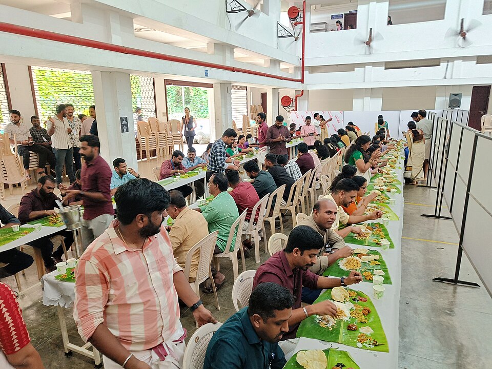 Onasadya at Jyothi Engineering College — an Onam Sadya organised as part of Onam celebrations at an engineering college, showing rows of students seated on the floor with banana leaves laid before them, servers moving along the rows with dishes, demonstrating how the Sadya tradition functions as a democratic communal institution where everyone — regardless of social position — receives the same feast on the same level
