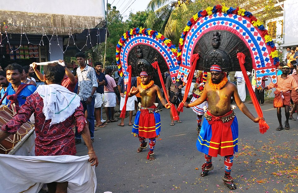 Poothan and Thira performers at the Deshamangalam Kottipparakkal Pooram festival in Kerala — the Poothan in full costume with imposing wooden headgear, peacock feathers, golden ornaments and the fierce protruding-tongue face mask, and the Thira wearing the black semi-circular Bhadrakali crown, both accompanied by Para and Thudi drum players during the annual Pooram festival